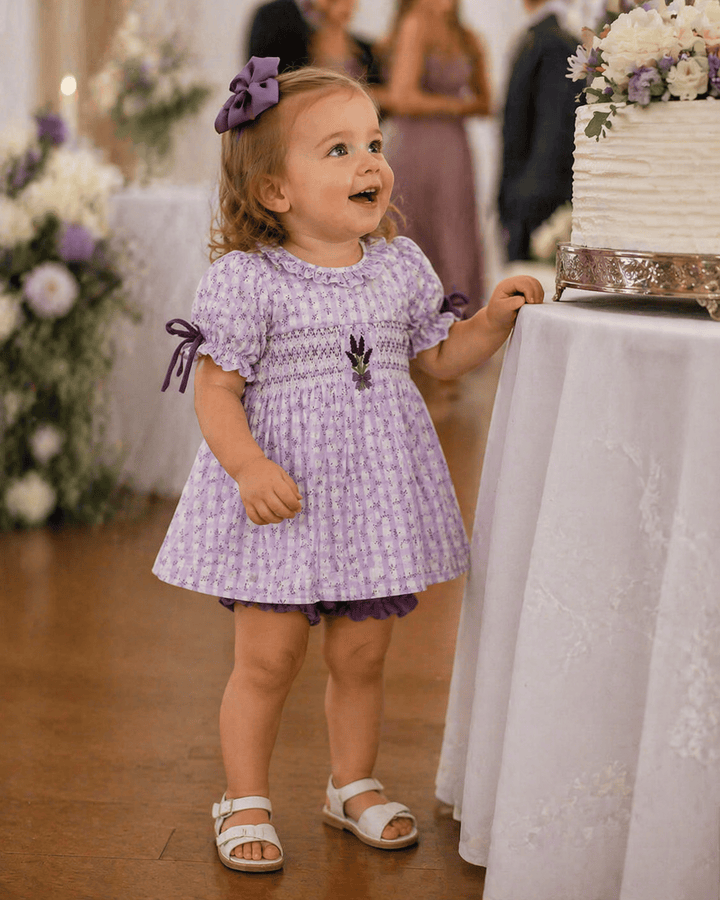 Child in a purple dress standing next to a wedding cake with floral decorations.