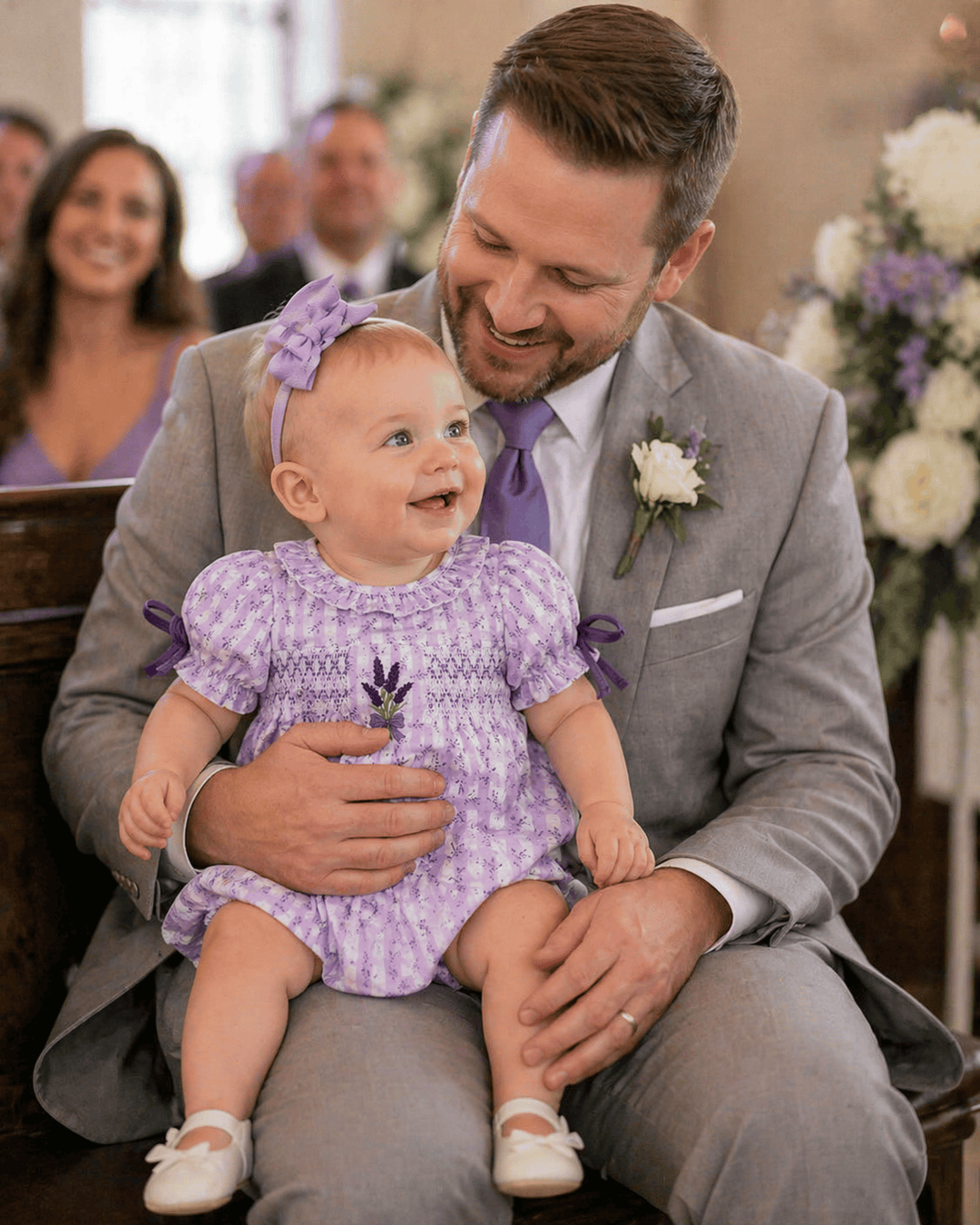 Man in a gray suit holding a baby in a purple dress with a floral arrangement in the background.
