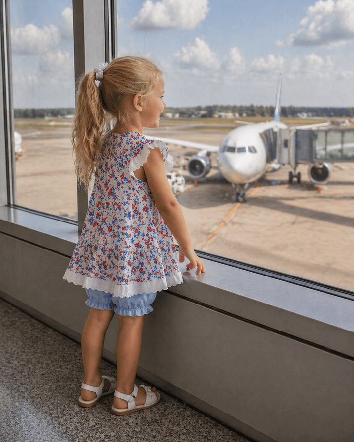 Child in a floral dress looking out of an airport window at an airplane.