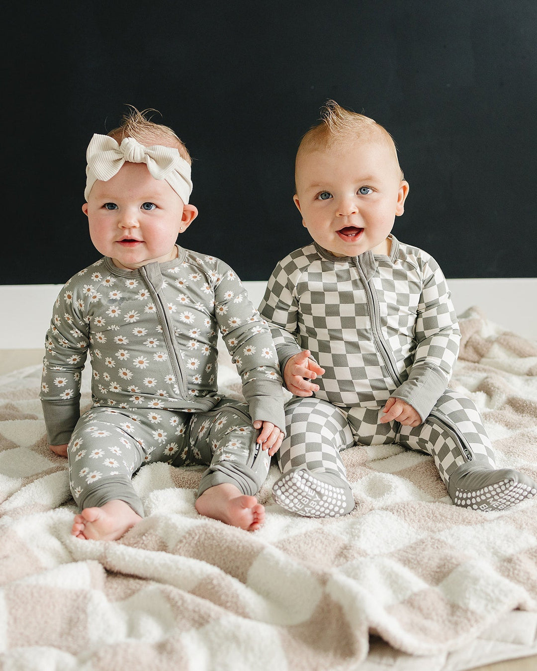 Two babies sitting on a blanket wearing patterned outfits against a black background