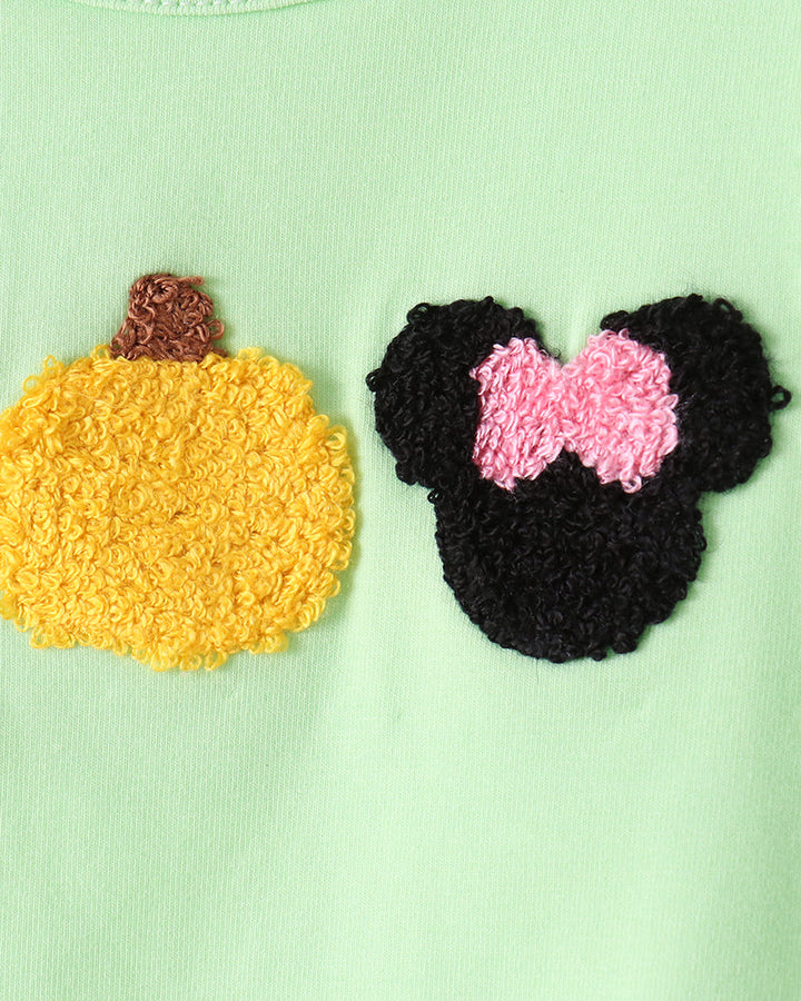 A close-up of a light mint green garment featuring two textured appliques: a yellow pumpkin with a brown stem on the left, and a black Minnie Mouse-style head silhouette with a pink bow on the right.