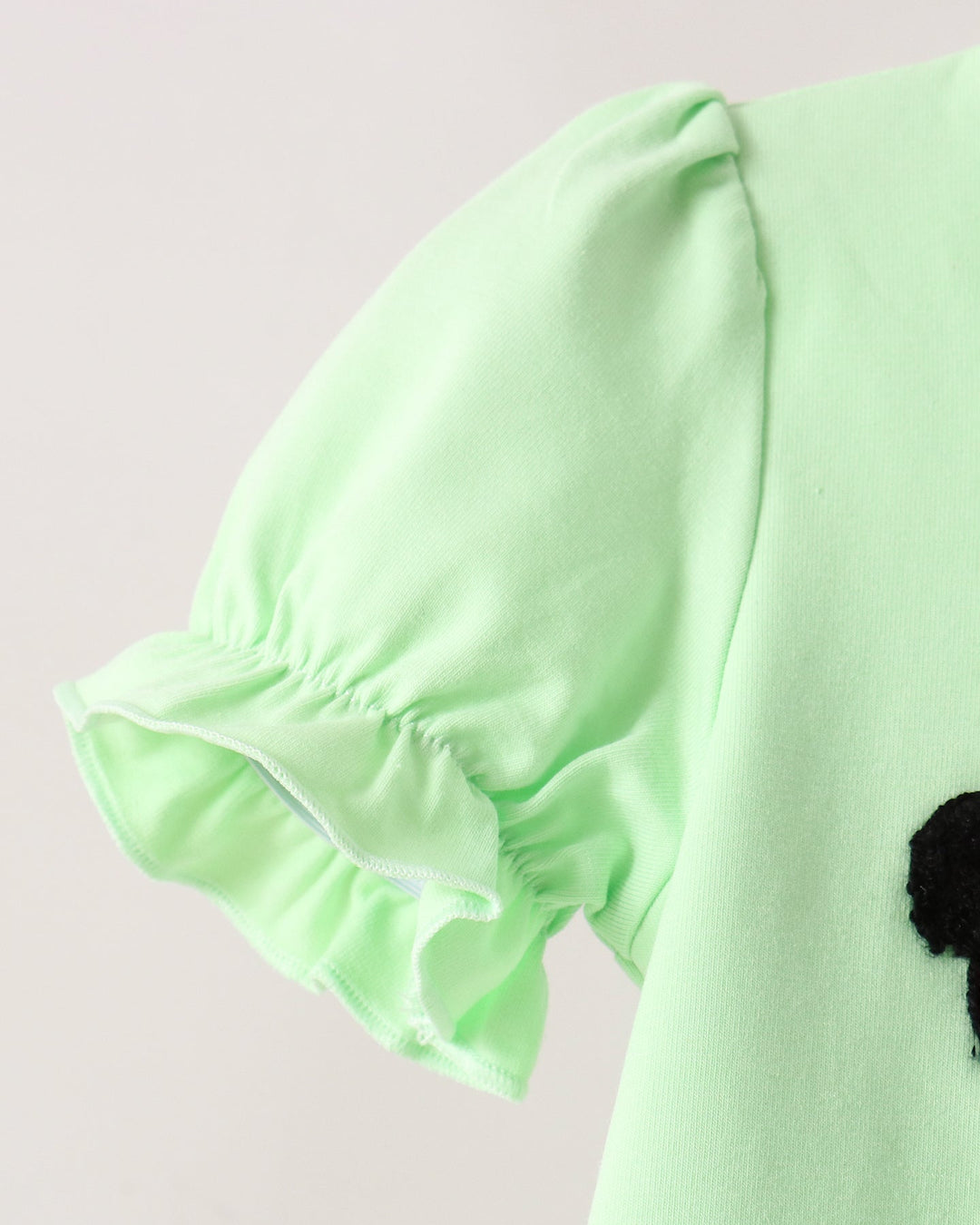 Close-up of a light mint green garment with a ruffled short sleeve and a small black pom-pom detail on the side, set against a plain light background.