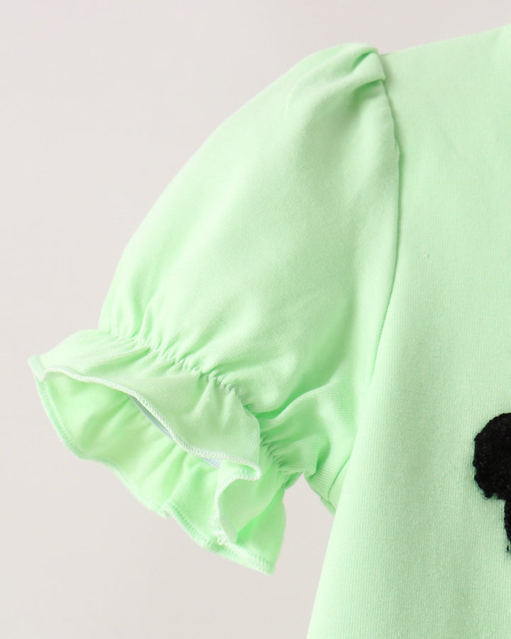 Close-up of a light mint green garment with a ruffled short sleeve and a small black pom-pom detail on the side, set against a plain light background.