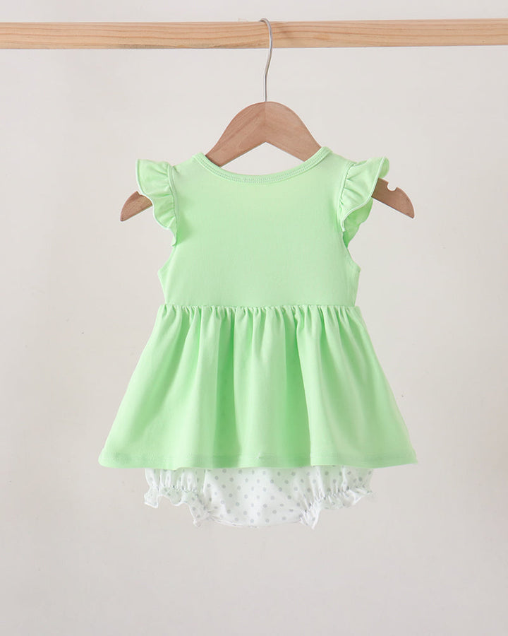 A light green sleeveless baby dress with ruffled cap sleeves and a gathered peplum, hanging on a wooden hanger against a plain light beige wall. A white ruffled underskirt with tiny polka dots peeks out from the hem.