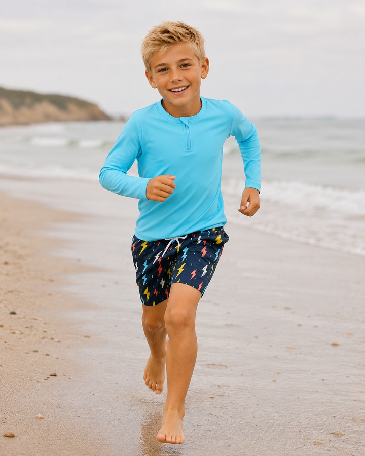Child running on a beach wearing a blue long-sleeve shirt and patterned shorts.