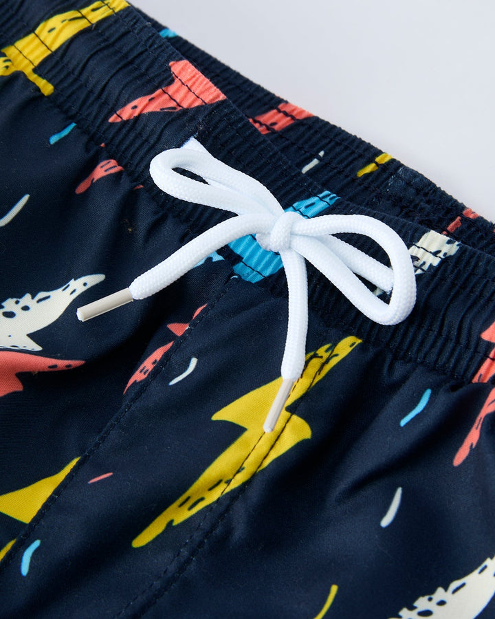 Navy swim shorts with colorful shark pattern and white drawstring on a light gray background