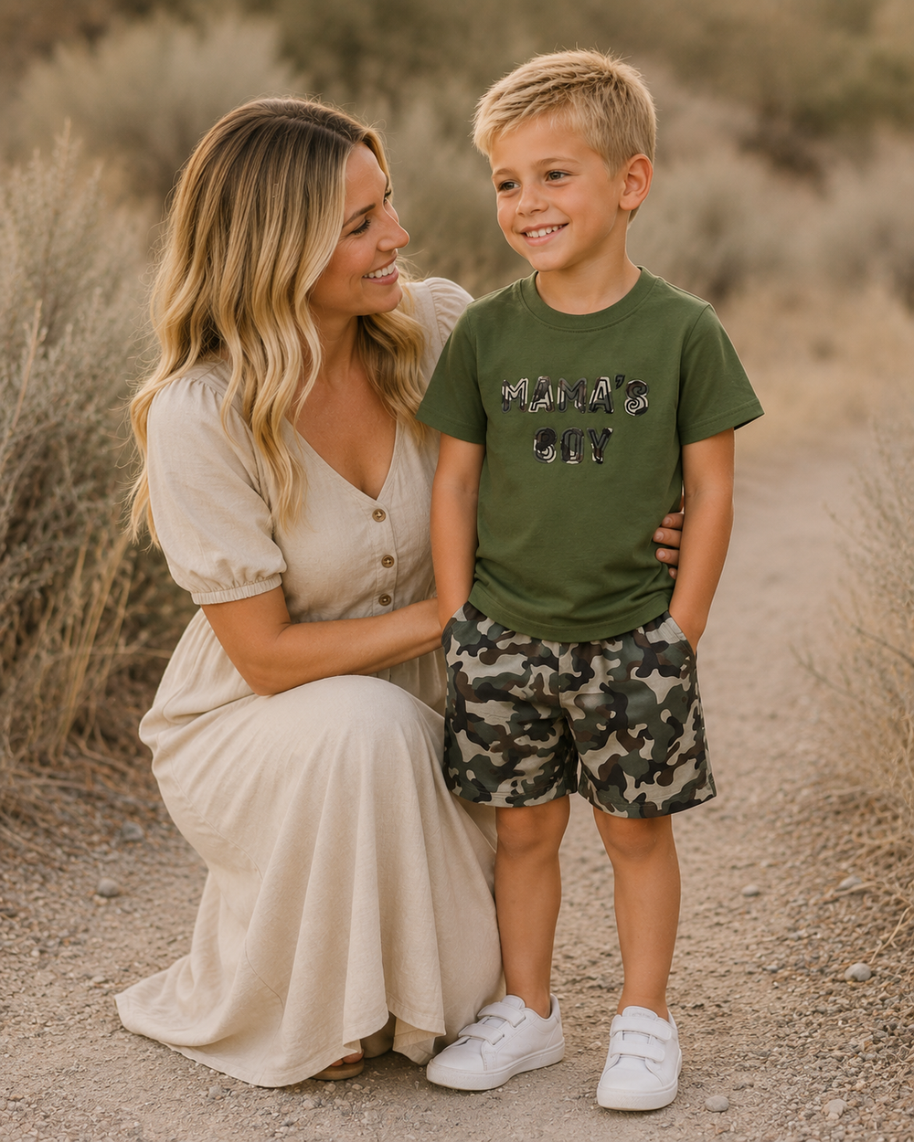 Woman in a long beige dress kneeling on a dirt path with a young boy in a green shirt and camouflage shorts.