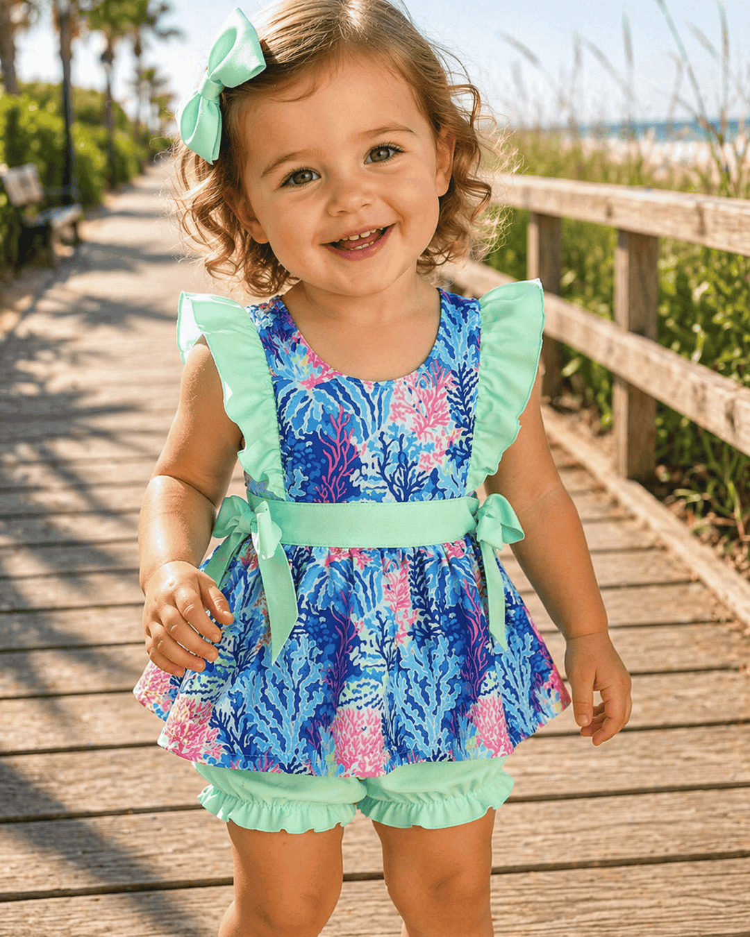 Child wearing a colorful dress with ruffles on a wooden boardwalk by the beach.