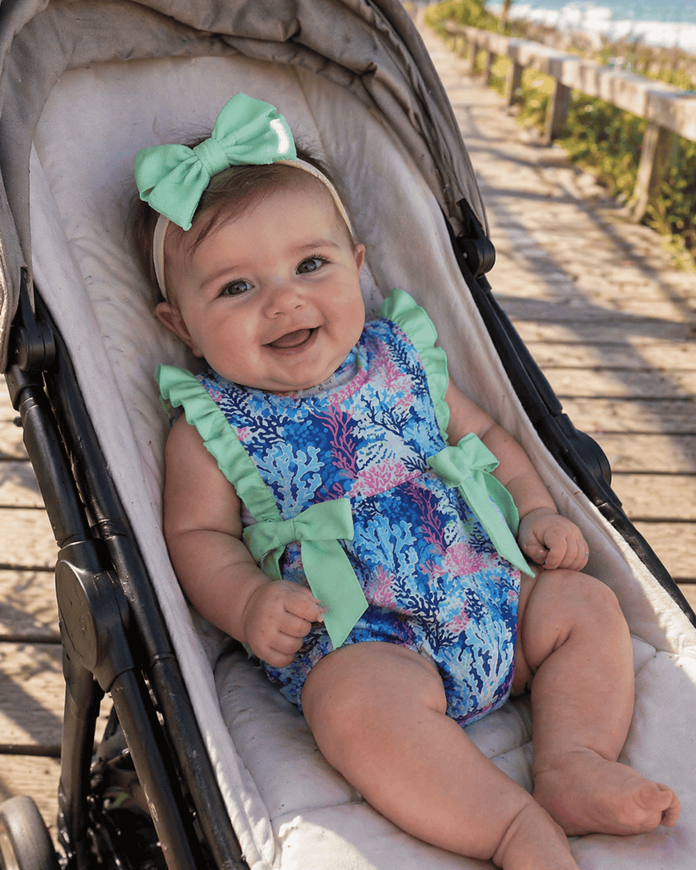 Baby in a colorful romper with green bows sitting in a stroller.