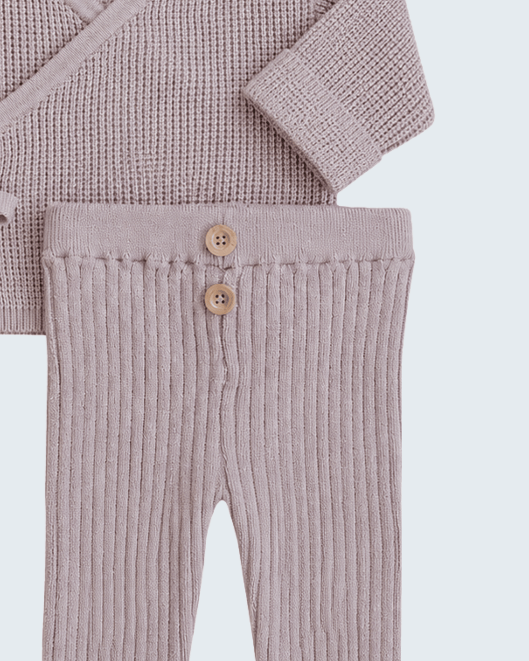 A close-up of a baby clothing set featuring ribbed knit pants in a dusty mauve-pink color with two wooden buttons, and a matching textured knit top partially visible at the top.