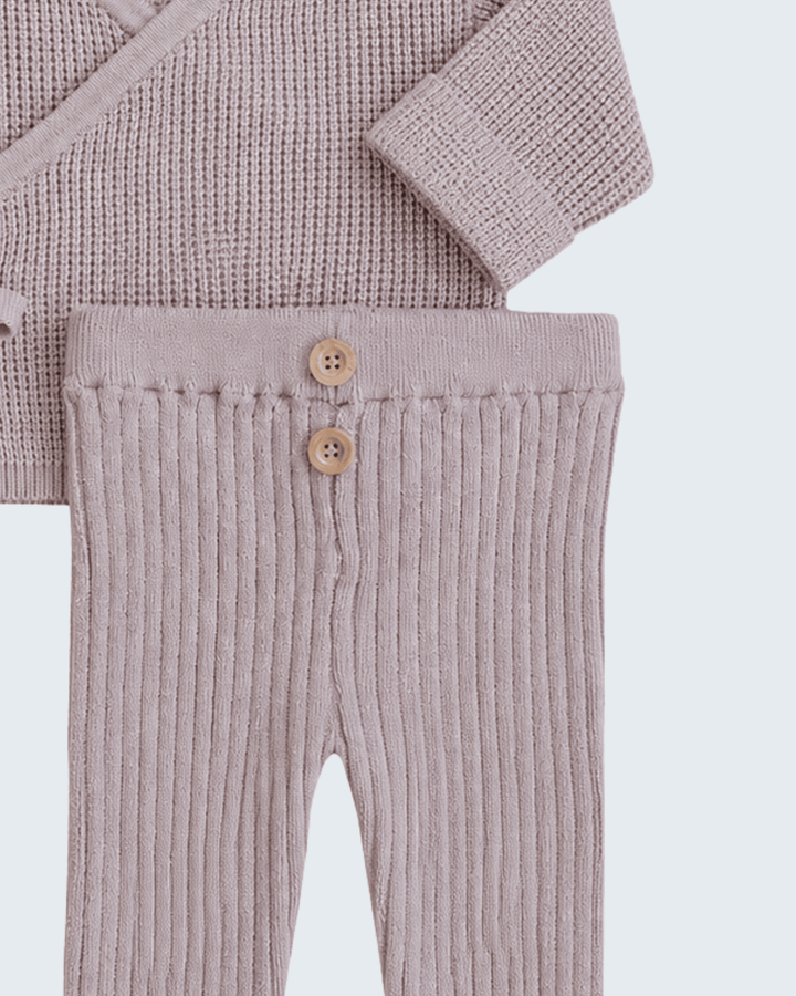 A close-up of a baby clothing set featuring ribbed knit pants in a dusty mauve-pink color with two wooden buttons, and a matching textured knit top partially visible at the top.