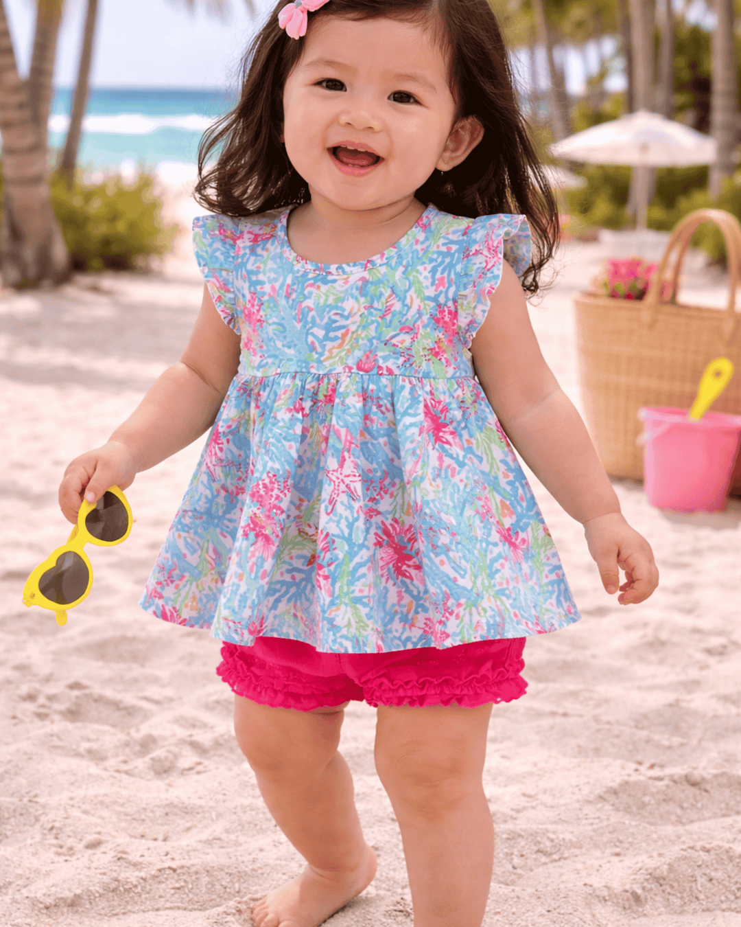 Child in a colorful dress on a beach with sunglasses and a basket in the background