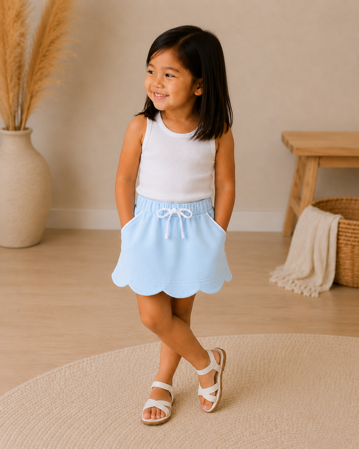 A young girl with dark hair stands in a brightly lit room wearing a sleeveless white top and a light blue skirt with a scalloped hem and white drawstring. She is wearing white sandals and standing on a round beige rug.