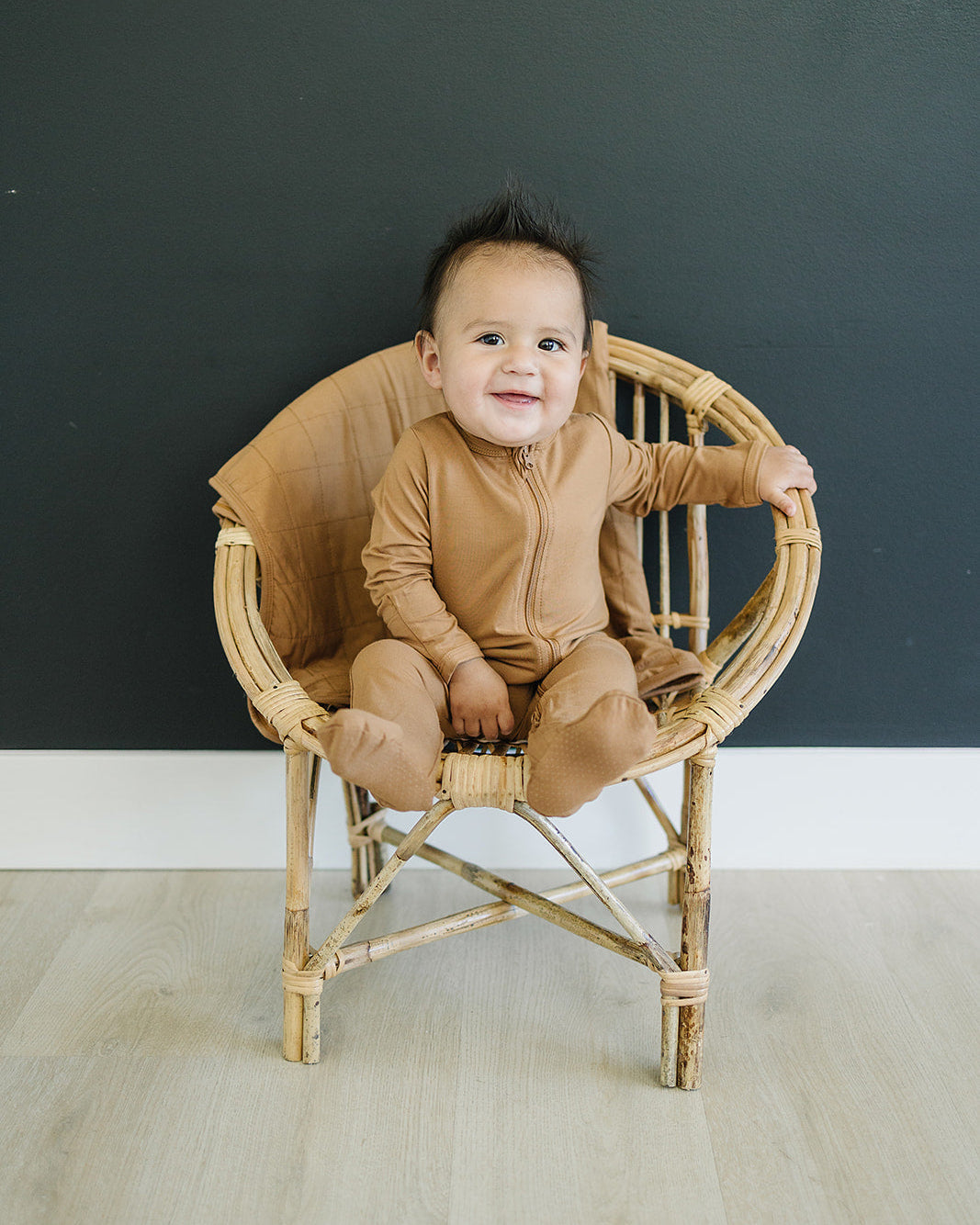 Baby in a brown outfit sitting on a wicker chair against a dark wall.