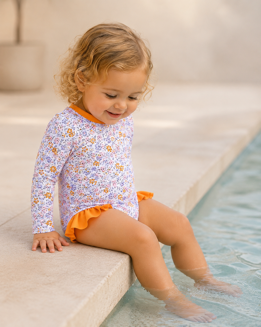 Child in a floral swimsuit with orange trim sitting by a pool.