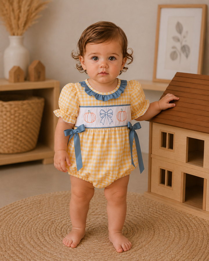 A toddler girl wearing a yellow and white gingham romper with blue ruffle trim, a white smocked chest panel with embroidered pumpkins and bows, and blue ribbon ties, standing in a styled nursery next to a wooden toy house.