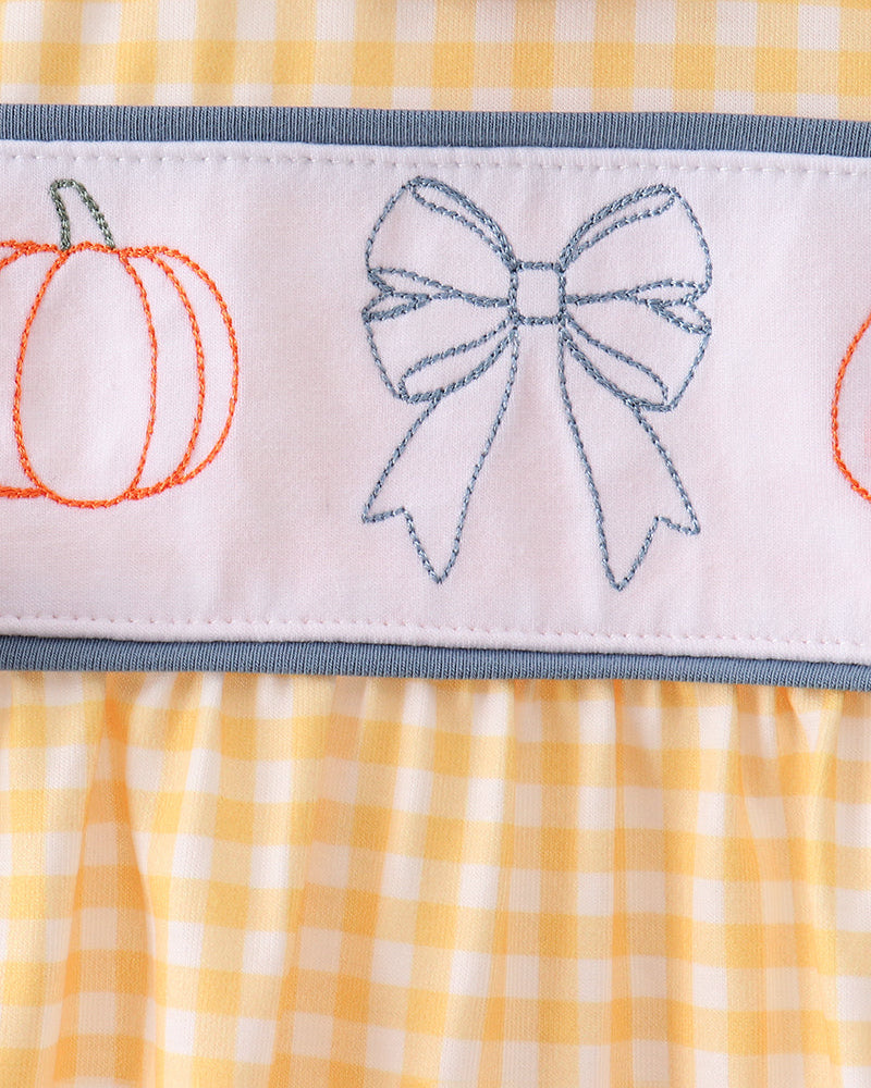 Close-up of a baby dress featuring a yellow and white gingham pattern, a light blue ruffled collar, and a white horizontal band with embroidered orange pumpkins and a blue bow.