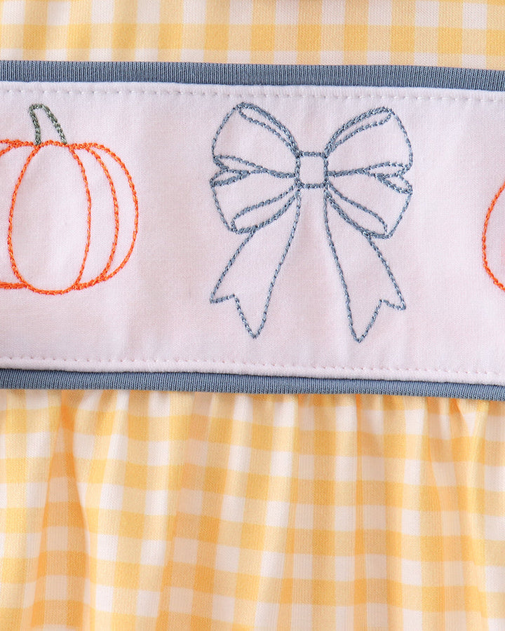 Close-up of a baby dress featuring a yellow and white gingham pattern, a light blue ruffled collar, and a white horizontal band with embroidered orange pumpkins and a blue bow.