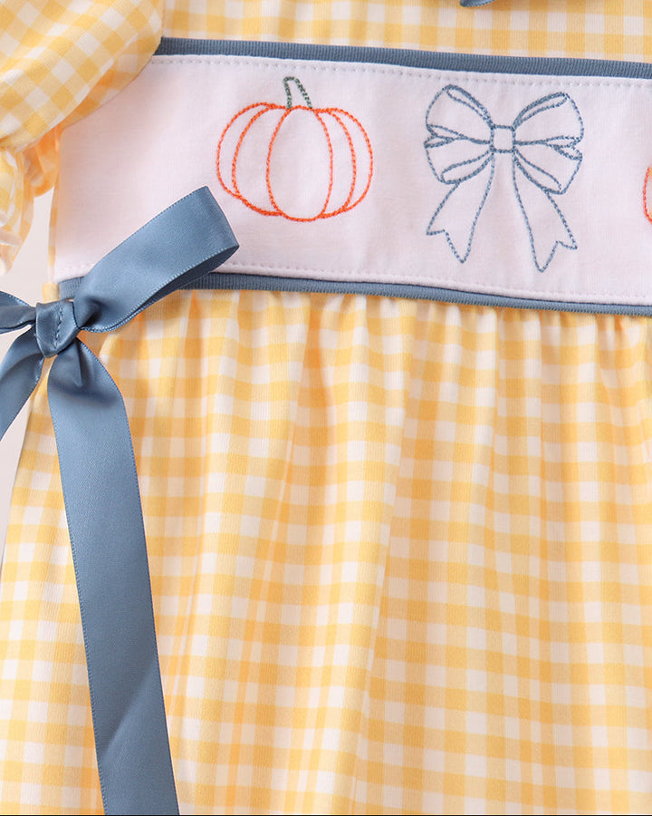 Close-up of a yellow and white gingham dress with a light pink waistband featuring embroidered orange pumpkin and blue bow designs, accented by a dusty blue ribbon and ruffled collar.