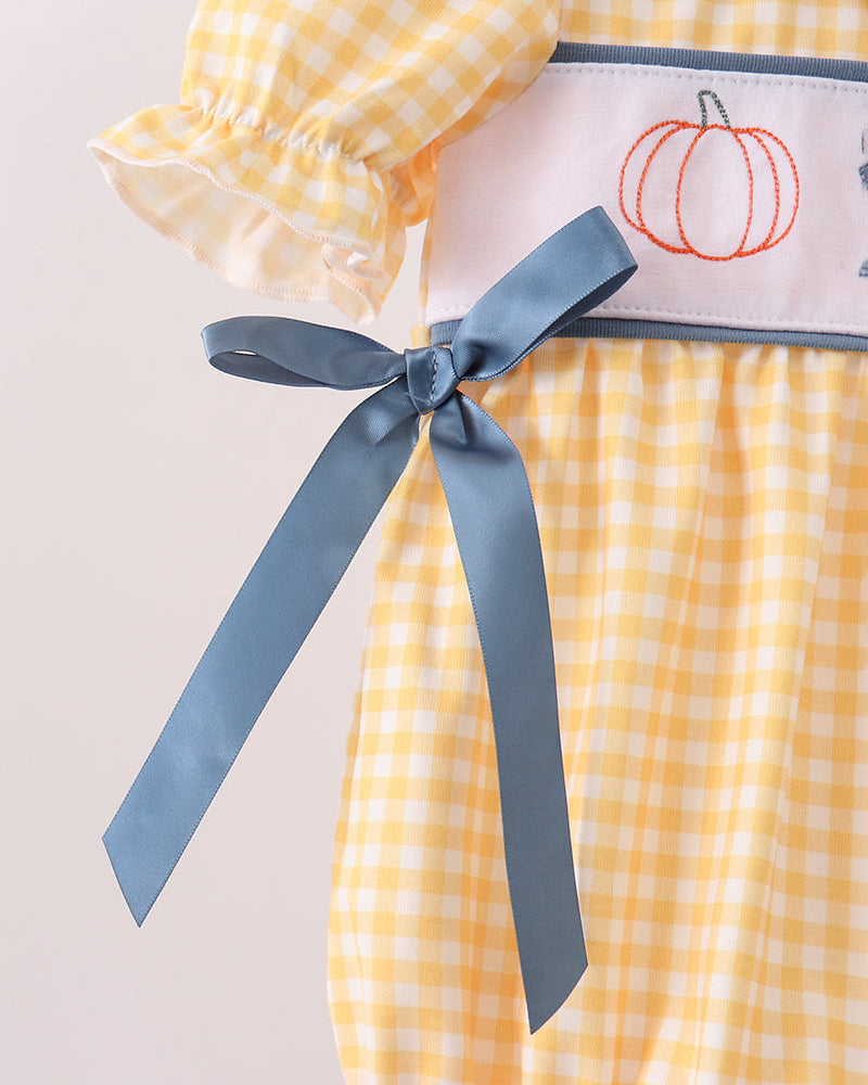 A close-up of a yellow and white gingham baby dress featuring a white waistband with an embroidered orange pumpkin and a dusty blue ribbon bow.