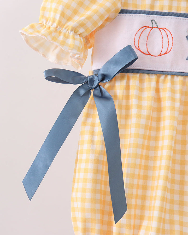 A close-up of a yellow and white gingham baby dress featuring a white waistband with an embroidered orange pumpkin and a dusty blue ribbon bow.