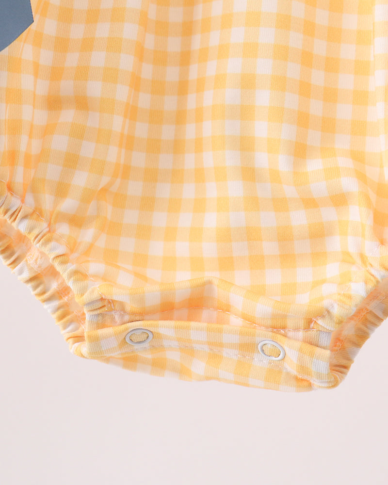 Close-up of a baby romper or bodysuit fabric in a yellow and white gingham check pattern, with elasticized leg openings and snap closures at the bottom, photographed against a plain light background.