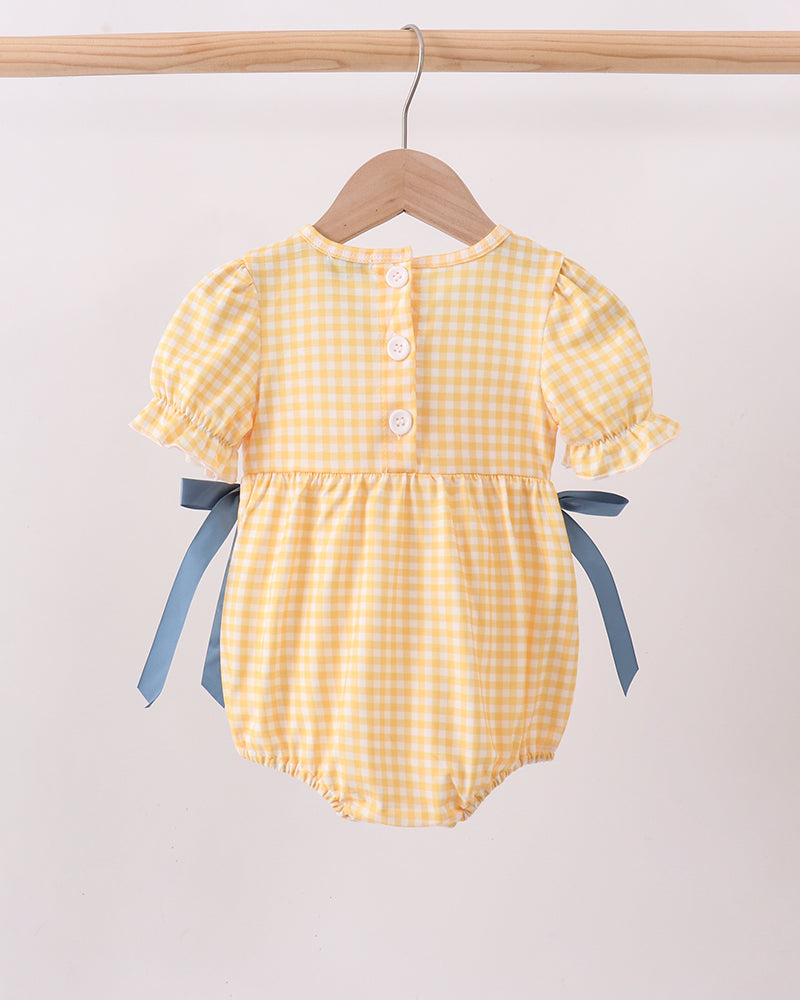 A yellow and white gingham baby romper with blue ribbon side bows, hanging on a wooden hanger from a light wood clothing rack against a plain off-white background.