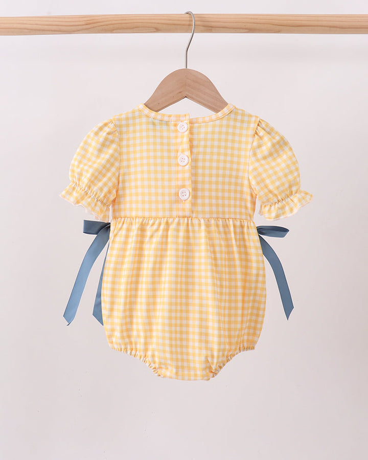 A yellow and white gingham baby romper with blue ribbon side bows, hanging on a wooden hanger from a light wood clothing rack against a plain off-white background.