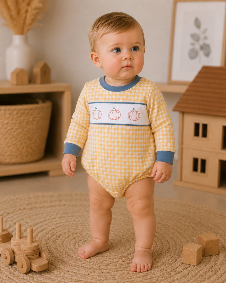 A baby standing on a woven rug wearing a yellow and white gingham long-sleeve bodysuit with a blue collar and cuffs, and a horizontal white stripe featuring three orange pumpkins. The background shows a cozy room with wooden toys and shelves.