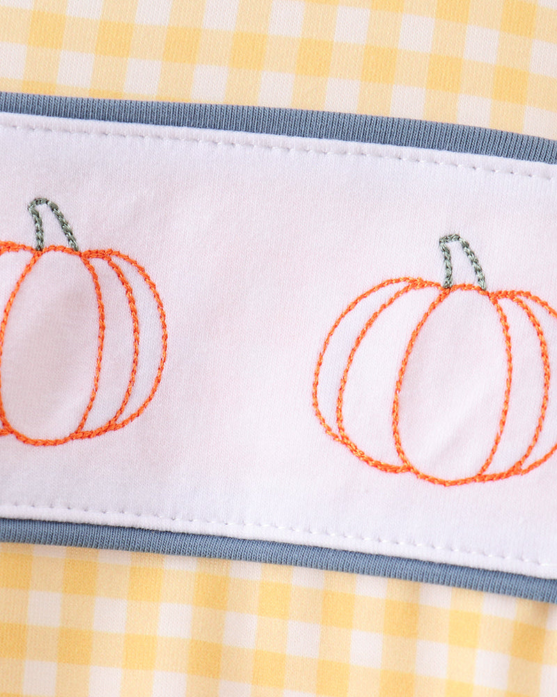 A close-up of a white fabric band with orange pumpkin embroidery and gray trim, laid over a yellow and white gingham background.