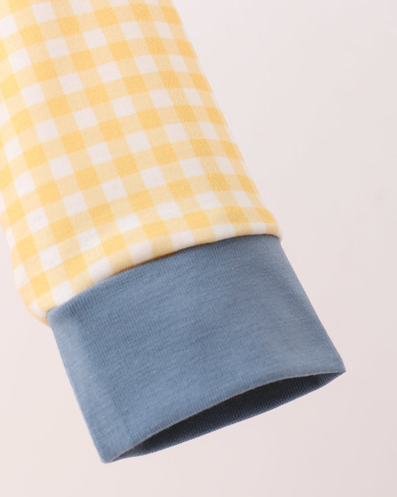 Close-up of a long-sleeve garment showing a yellow and white gingham pattern on the upper section and a solid dusty blue ribbed cuff on the lower section, set against a plain light beige background.