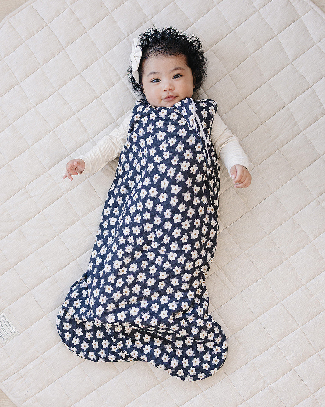 Baby lying on a white quilted surface wearing a navy blue floral sleep bag.