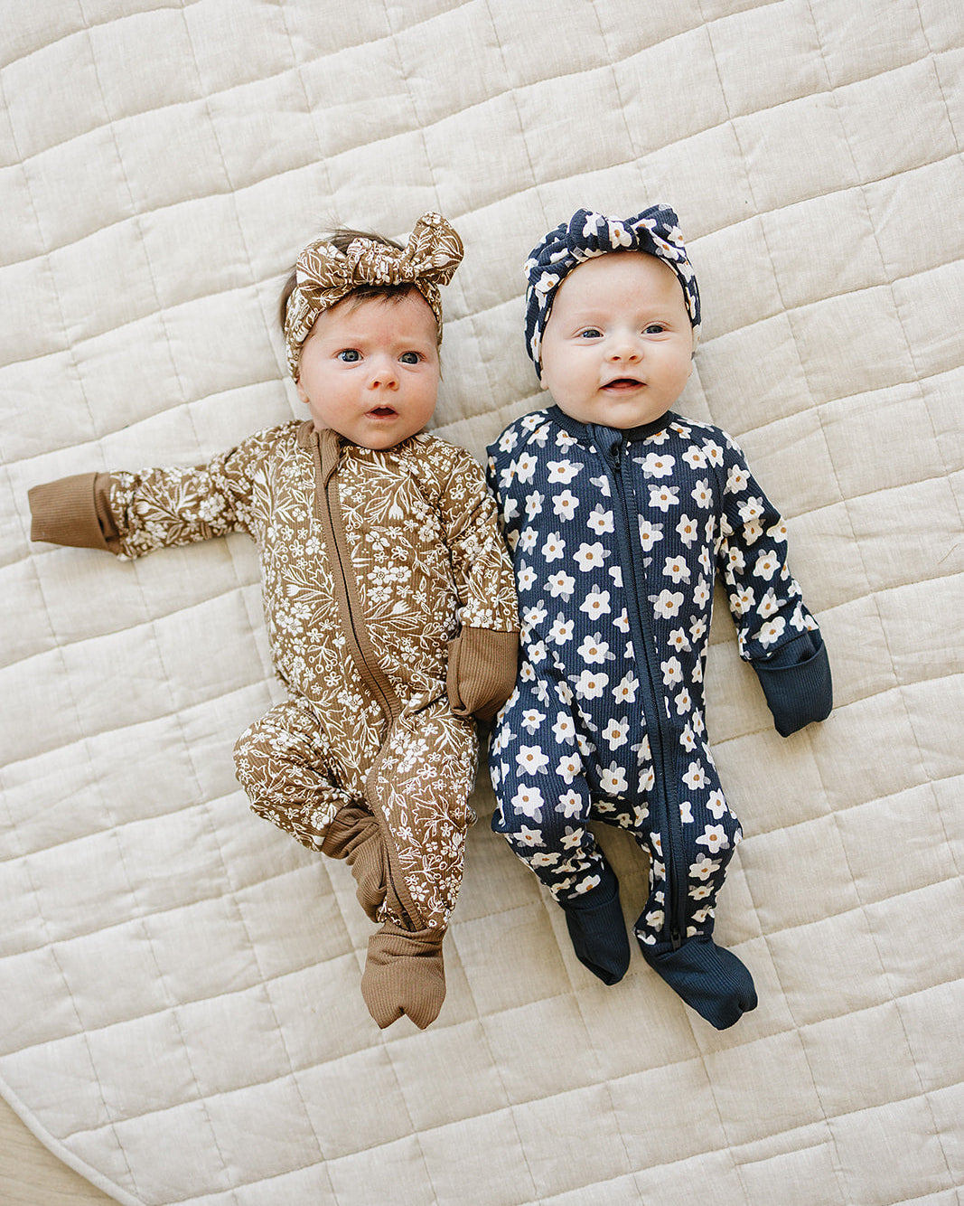 Two babies wearing patterned outfits lying on a quilted surface.