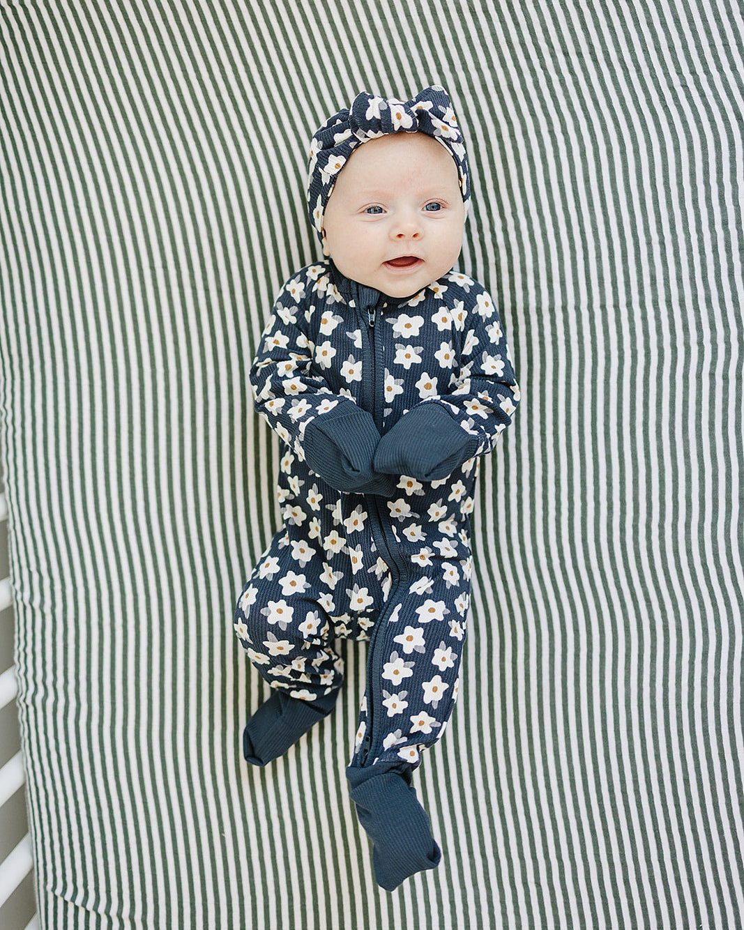 Baby in a floral onesie and navy hat lying on a striped fabric background