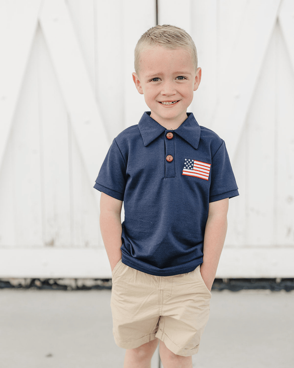 Young boy wearing a navy polo shirt with an American flag patch and beige shorts, standing against a white curtain background.