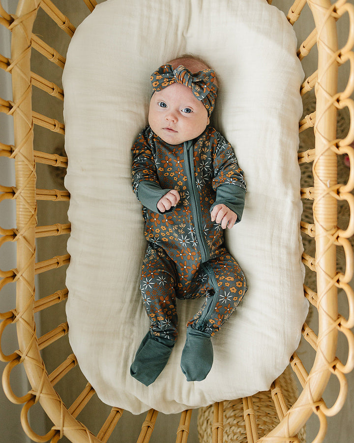 Baby in a patterned outfit lying on a white cushion inside a wicker crib.