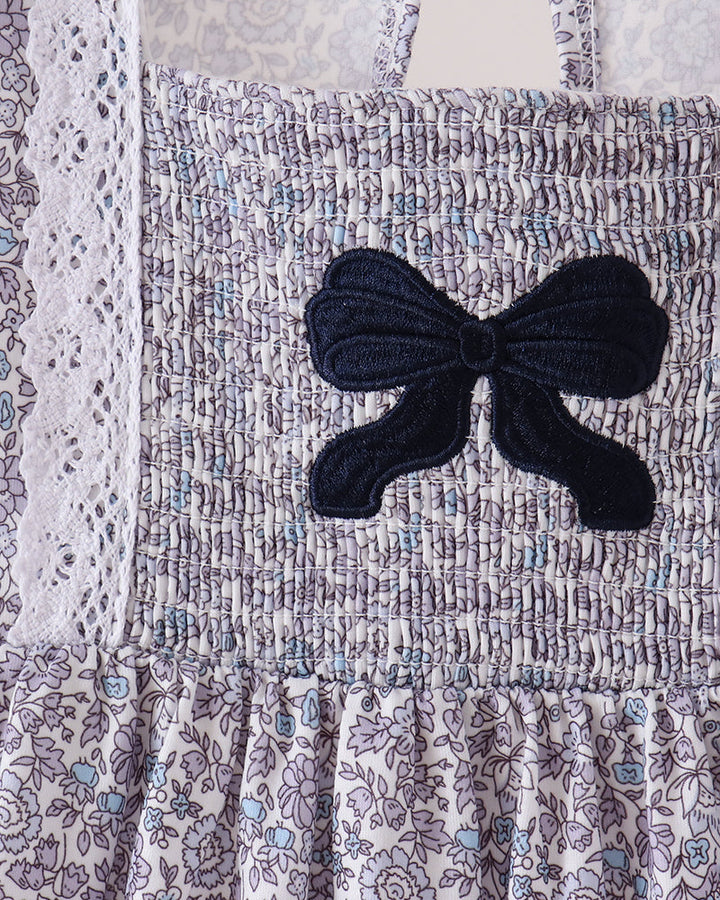 Close-up of a smocked dress or bodice with a floral print in lavender, blue, and white, featuring a large dark navy bow applique on the front.