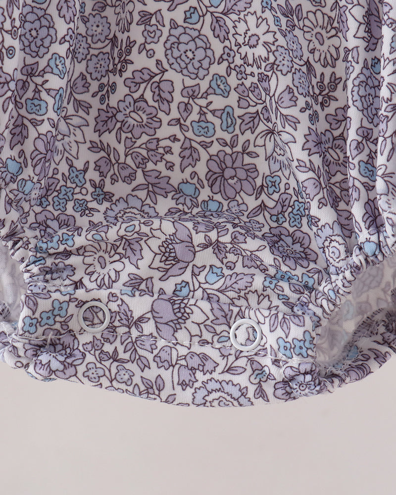 Close-up of a floral-patterned fabric garment, likely a baby bloomer or diaper cover, featuring a white base with purple and light blue floral print and snap closures at the bottom.