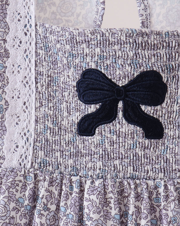 Close-up of a smocked dress or bodice with a floral print in lavender, blue, and white, featuring a large dark navy bow applique on the front.