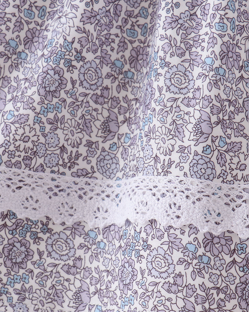 Close-up of a floral fabric with a lavender and light blue print on a white background, featuring a decorative white lace trim.