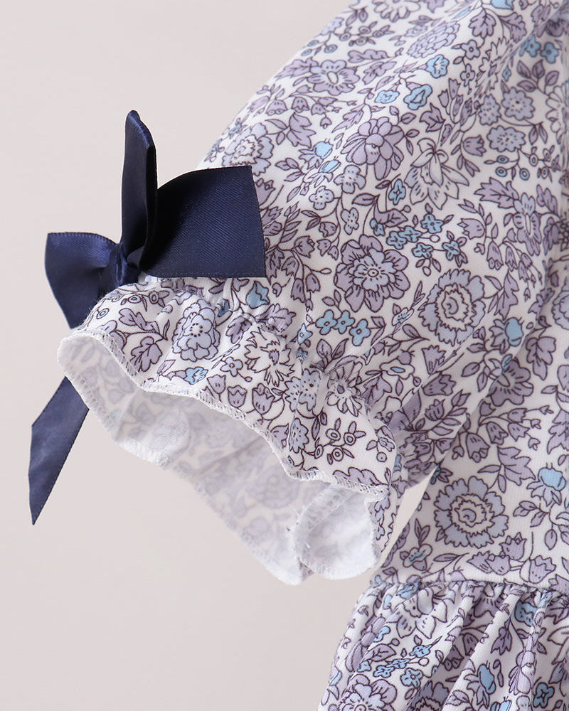 A close-up of a floral dress or garment featuring a white base with a purple and light blue floral pattern, a navy blue bow on the sleeve, and a ruffled lace trim.