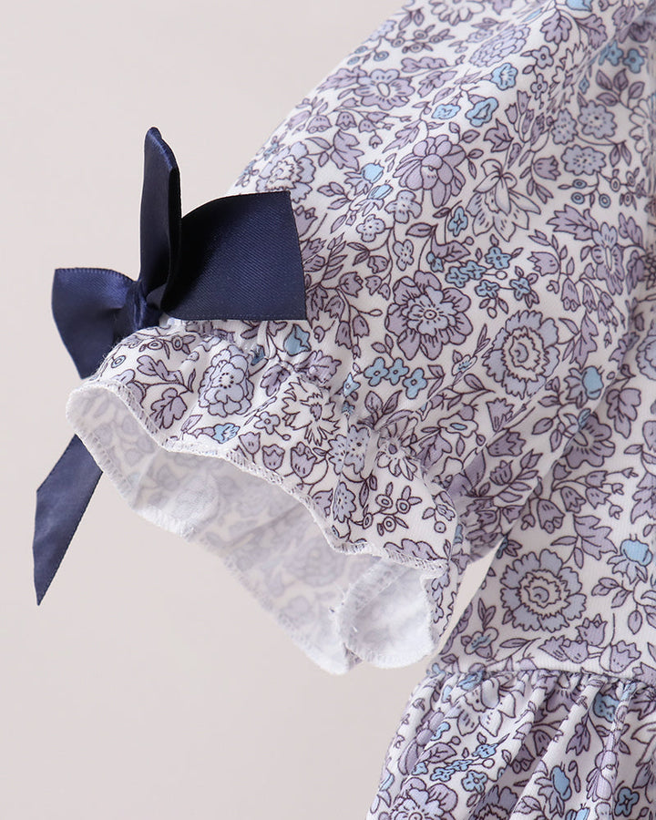 A close-up of a floral dress or garment featuring a white base with a purple and light blue floral pattern, a navy blue bow on the sleeve, and a ruffled lace trim.