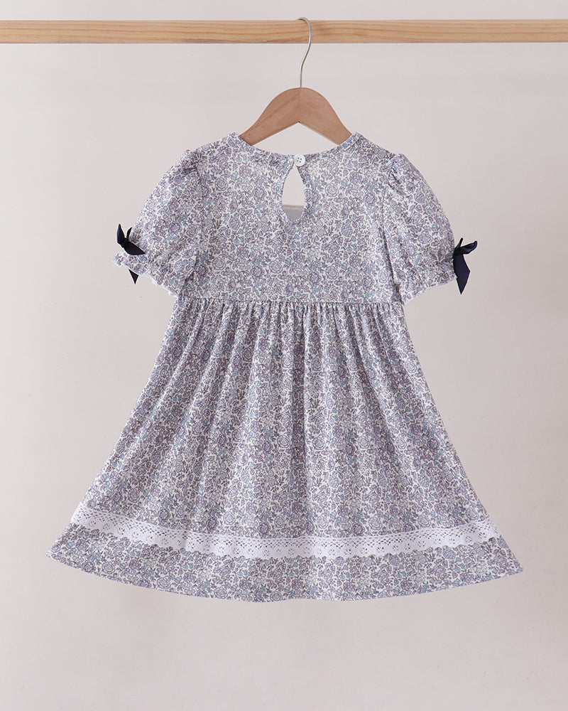A short-sleeve baby dress with a silver and white floral pattern, hanging on a wooden hanger against a plain beige wall. The dress features puff sleeves with black ribbon bows at the cuffs, a smocked bodice, and a lace-trimmed hem.