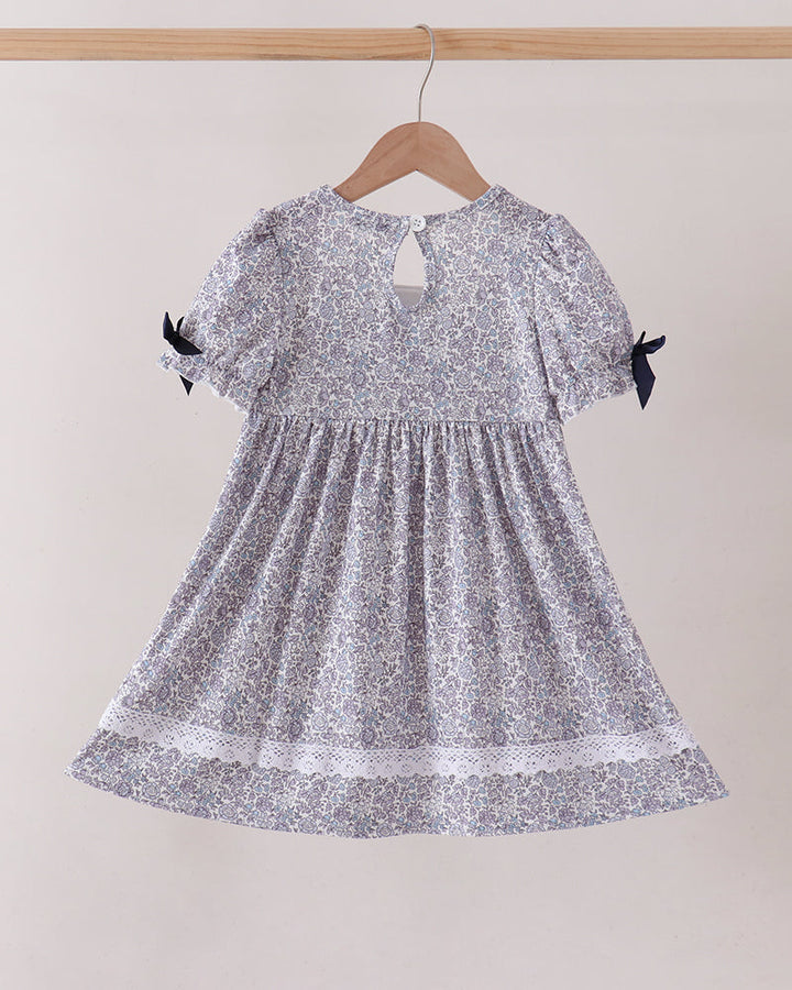 A short-sleeve baby dress with a silver and white floral pattern, hanging on a wooden hanger against a plain beige wall. The dress features puff sleeves with black ribbon bows at the cuffs, a smocked bodice, and a lace-trimmed hem.