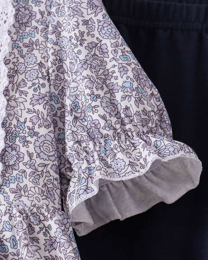 Close-up of a floral-patterned blouse with a ruffled short sleeve, featuring a white base with purple and light blue floral print, shown next to dark navy shorts.