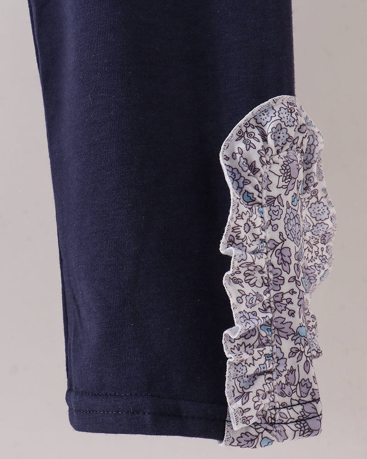 A close-up of a navy blue pant, featuring a gathered ruffle of floral-patterned fabric attached near the lower side. The floral pattern is white with purple and blue details.