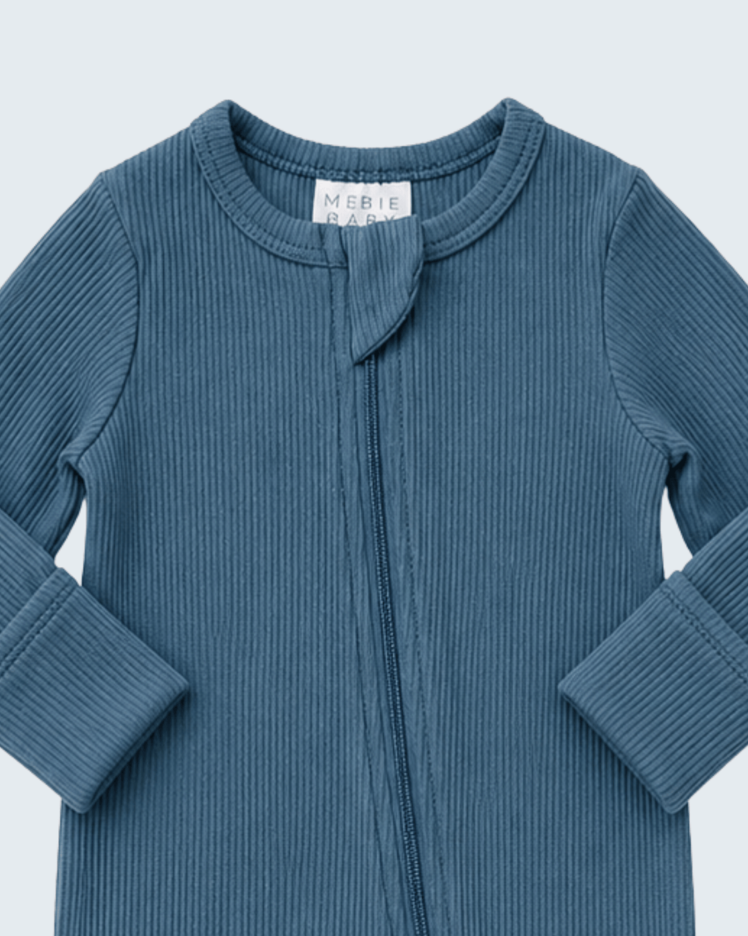 A close-up of a ribbed knit baby top in a dusty slate blue color, featuring a front zipper and a small fabric bow detail at the neckline. The brand label &