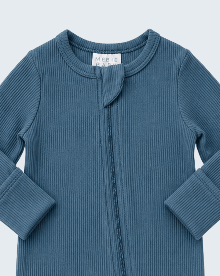 A close-up of a ribbed knit baby top in a dusty slate blue color, featuring a front zipper and a small fabric bow detail at the neckline. The brand label &
