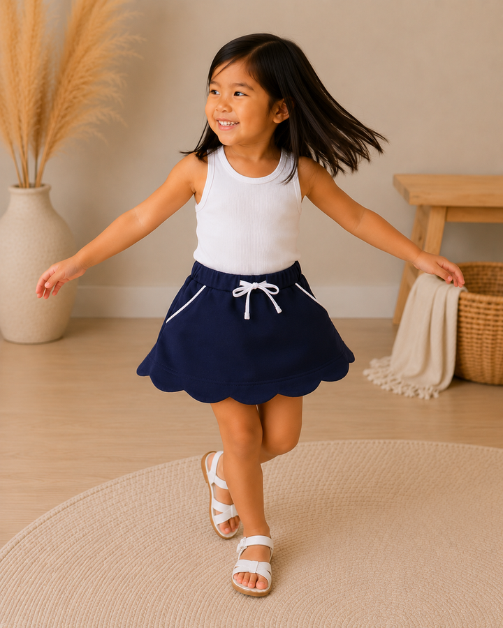 A young girl with dark hair is smiling and twirling while wearing a white sleeveless tank top and a navy blue skort with white drawstring details. She is standing on a round beige rug in a warm, neutral-toned room with a wooden bench, woven basket, and pampas grass in a vase.