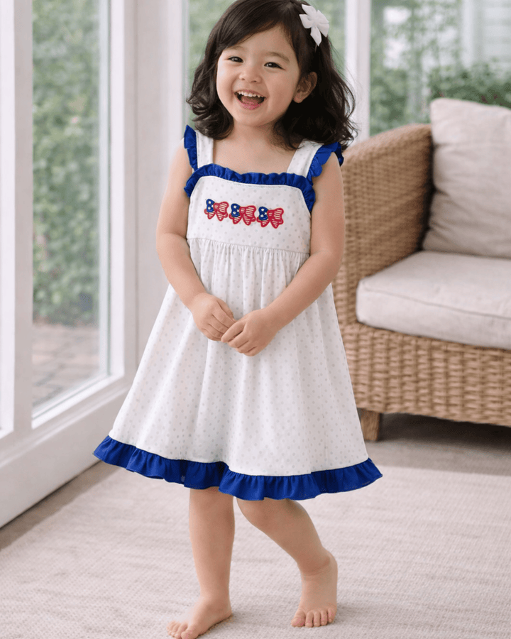 Young girl in a white dress with blue trim and red accents standing in a room with large windows.